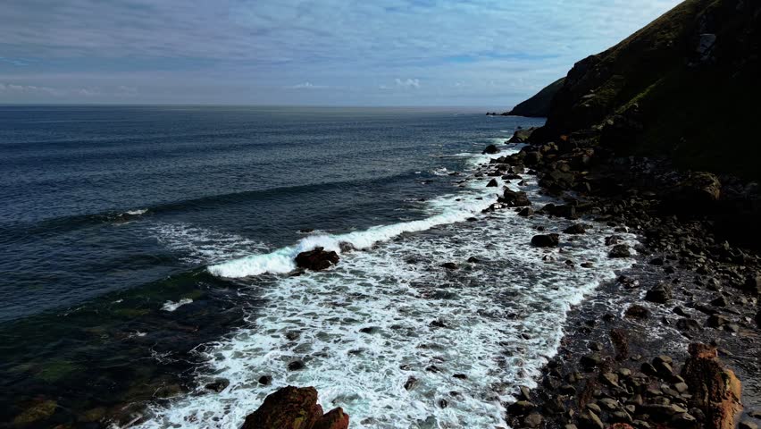 Waves crashing onto rugged coastline of North Sea in Scotland. Swells pounding dark rocks along steep shore. Foam swirling around jagged stones near grassy cliffs. Tides battering coastal boulders