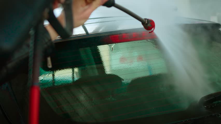 Unrecognizable woman spraying rear window of car with high-pressure water at self-service wash. Female rinsing back glass using strong water stream. Person cleaning rear windshield of vehicle with
