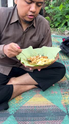 Portrait of Asian man almost finished a healthy dish of rice with tempeh and sauce on a teak leaf outdoors. Ideal for food blog or culinary promotion. sstkTravel.