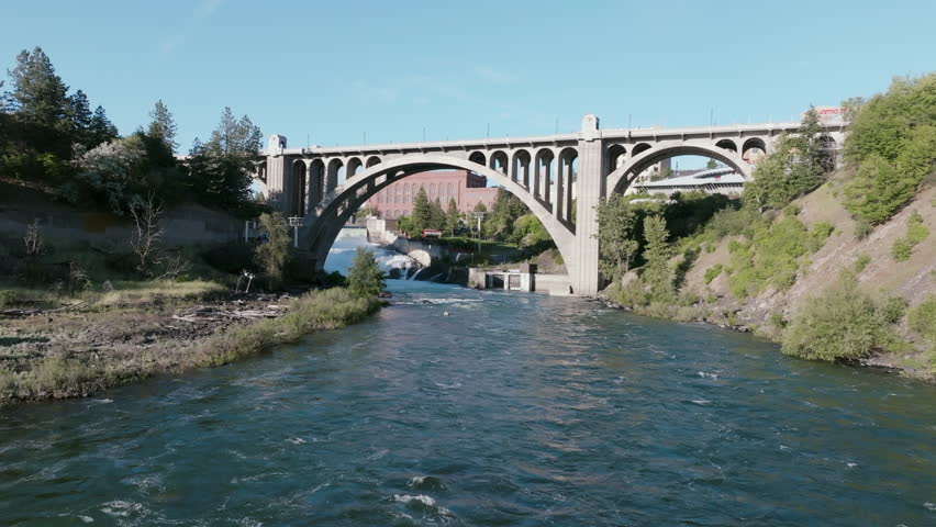 A downstream view of the Spokane River rushing beneath the Monroe Street Bridge with Spokane’s redbrick skyline and lush green banks on both sides.