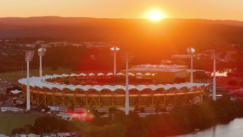 Drone glides over a large sports stadium at sunset, capturing dramatic lighting, illuminated field lights, and the adjacent river in Gold Coast, Australia