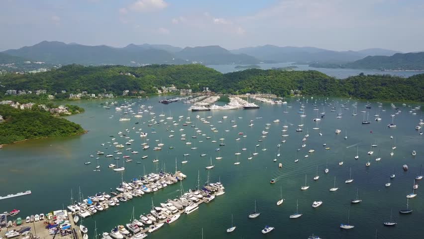 Drone video captures Discovery Bay marina in Hong Kong, showcasing hundreds of anchored boats against lush green hills and tranquil waters under a bright blue sky. Scenic beauty.
