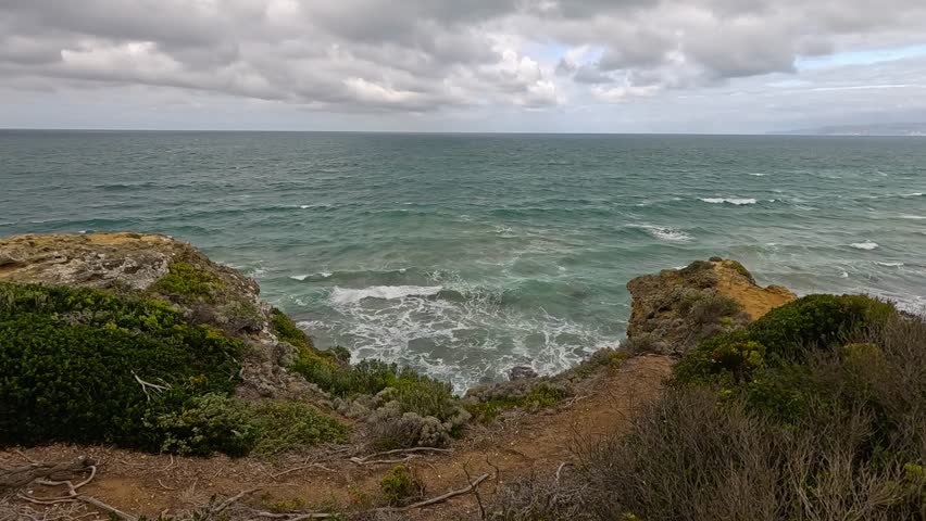 Waves surge against rugged cliffs and coastal vegetation under cloudy skies. Static wide shot captures dynamic ocean movement and dramatic natural landscape along Great Ocean Road - Powered by Shutterstock - Get 15% off with code: PIKWIZARD15