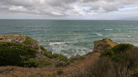 Waves surge against rugged cliffs and coastal vegetation under cloudy skies. Static wide shot captures dynamic ocean movement and dramatic natural landscape along Great Ocean Road - Powered by Shutterstock - Get 15% off with code: PIKWIZARD15