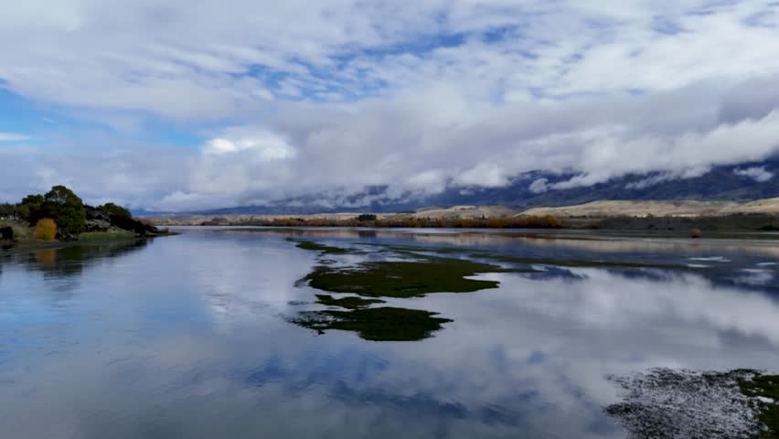 Drone glides above tranquil wetlands with water reflections, lush greenery, and distant mountains under dramatic clouds. Smooth forward camera movement, natural daylight