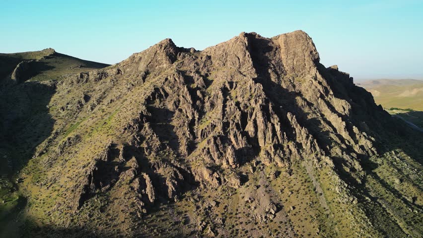 Steep rocky slopes stretch across the Uzbek landscape. Sunlight reveals ancient layers. Green vegetation dots the mountainside. Peaks rise sharply against the sky.