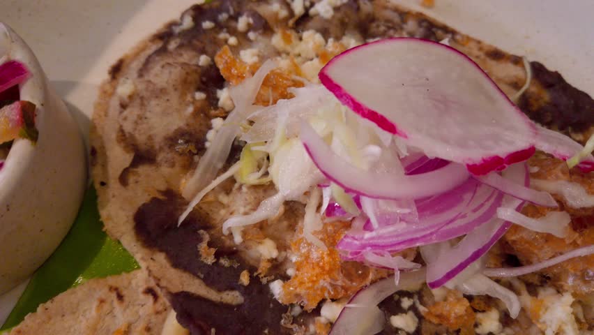 Close-up of a savory Oaxacan tlayuda layered with black beans, cheese, fried pork skin, radish, pickled onions and lettuce, served with a side of spicy salsa