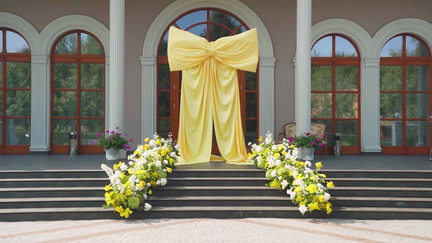 Beautiful wedding arch, huge yellow bow with yellow and white fresh flowers on the steps of a building in a classic style. Wedding arch on a bright sunny day without people before wedding ceremony.
