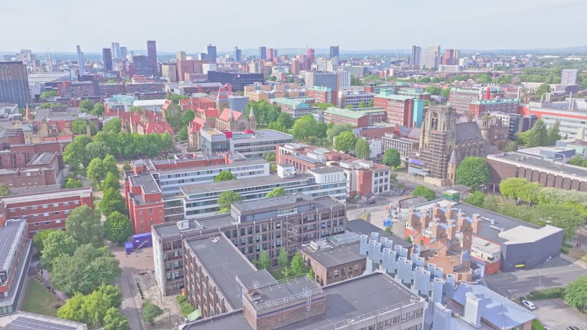 Drone ascends from the University of Manchester campus, showcasing red-brick buildings, the Whitworth Hall spire, and the city