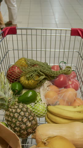 Vertical UGC shot of grocery cart with fruits and vegetables being pushed by unknown man during shopping in supermarket