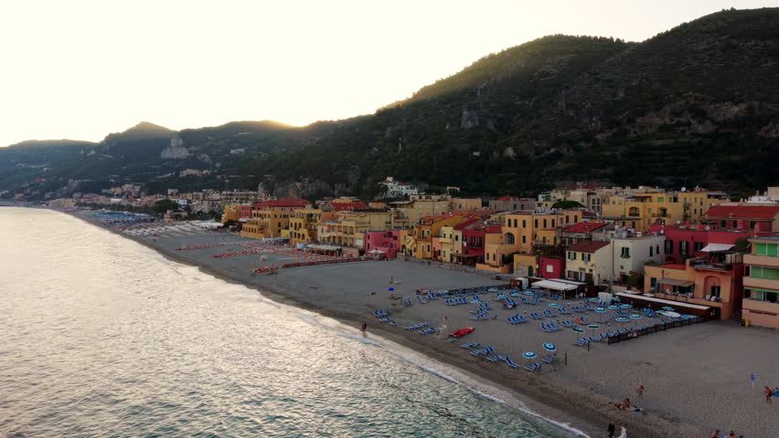 Aerial drone shot pulling back over the sea, revealing the colorful town of Varigotti, the beach, and the hills behind, with a seagull flying across at sunset.