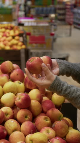 Vertical view of hands of unrecognisable woman wearing grey sweater choosing apples in supermarket