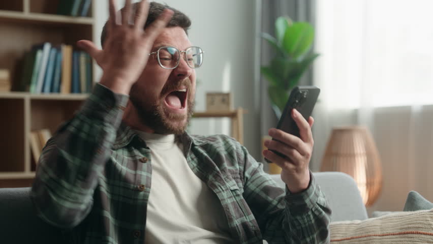 Caucasian adult man male guy at home living room nervous looking at mobile phone online stream watching football sport game betting casino waiting hoping lost upset failure bad result smartphone app