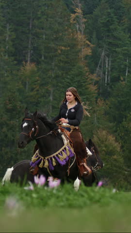 Vertical video of Confident young woman, a cowgirl with spirit of freedom and adventure riding a majestic black horse, through breathtaking mountain terrain, slow motion shot.