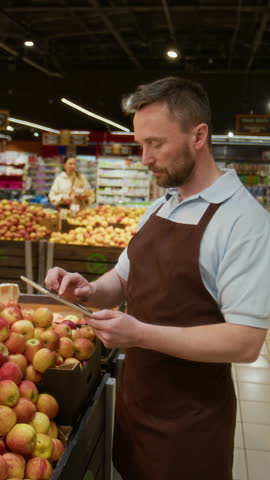Vertical view of Caucasian male supermarket worker examining apples for freshness and keeping notes on digital tablet
