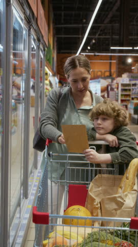 Vertical view of gen alpha kid holding piece of paper with shopping list, mother reading it, opening sliding door of refrigerator, taking plastic milk container from shelf and putting it in cart