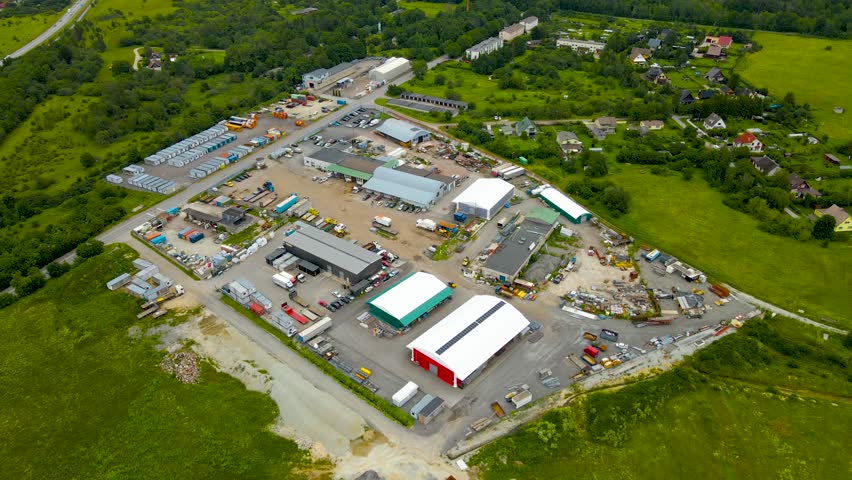 High altitude aerial drone footage orbiting around a countryside car park or an idustrial area that is fenced off from a highway and summwe green nature around it. Big warehouses and rooftops visible.