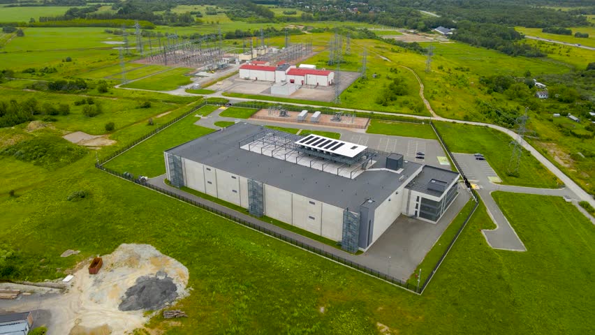 Aerial drone footage of a large and modern data centre or server housing complex warehouse during a cloudy day in europe countryside. A large electrical station in the background with electrical lines