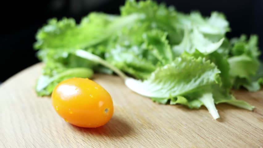 Chef cuts a tomato in half on a wooden board on a black background. Close-up of tomatoes and lettuce. Concept of healthy eating and organic farm products. 4k footage.