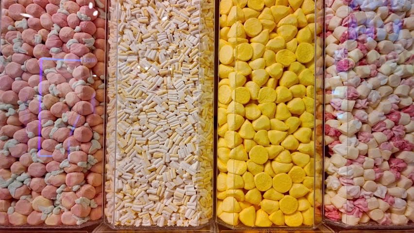 Close-up view of colorful candies neatly sorted into four vertical transparent containers. The sweets include pink, yellow, white, and mixed pastel colors, creating a vibrant and inviting visual