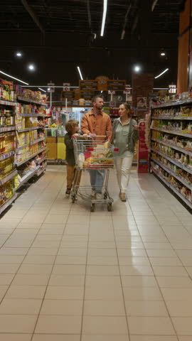 Vertical view of Caucasian married couple and their child walking down aisle of supermarket pushing cart with groceries and chatting