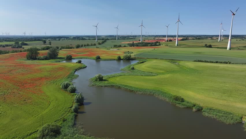 Aerial view of vibrant red poppy fields and wind turbines contrasting with lush green fields and tranquil water, Brussow Uckermark, Brandenburg, Germany.