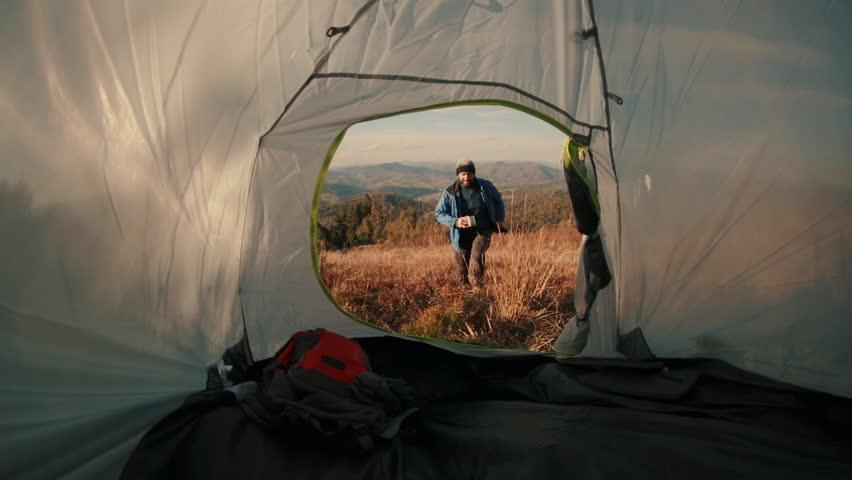 A male traveler runs into his tent set on a scenic mountain slope. Filmed from inside the tent, the breathtaking view opens behind him, capturing the thrill of adventure and natural beauty at sunset.