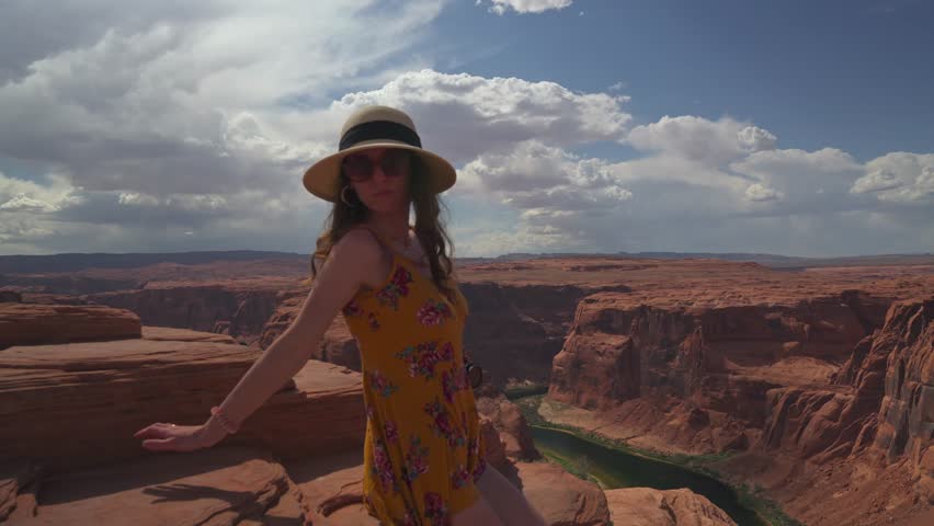oung woman in a summer dress posing at the edge of Horseshoe Bend canyon with dramatic clouds and the Colorado River far below.
