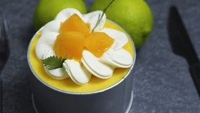 Small mango mousse cake decorated with whipped cream rosettes, mango cubes, and mint leaves, sitting on a dark gray textured surface with two limes and a knife in the background - Powered by Shutterstock - Get 15% off with code: PIKWIZARD15