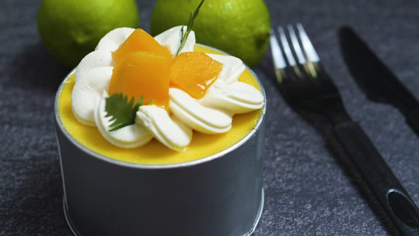 Small mango mousse cake decorated with whipped cream, peach slices, and a sprig of mint, served on a dark textured surface with limes in the background and a fork and knife ready for use