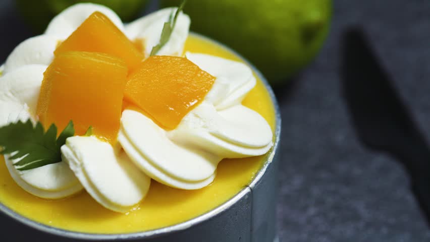 Small mango mousse cake decorated with whipped cream, peach slices and parsley leaves, sitting on a dark gray slate table with a blurred lime in the background