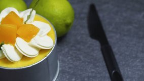 Small mango mousse cake decorated with whipped cream rosettes, mango cubes, and a sprig of mint, sitting on a dark gray surface with two limes in the background - Powered by Shutterstock - Get 15% off with code: PIKWIZARD15