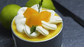 Small mango mousse cake decorated with whipped cream rosettes, mango slices, and fresh herbs, sitting on a dark gray surface with whole limes in the background - Powered by Shutterstock - Get 15% off with code: PIKWIZARD15