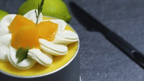 Small mango mousse cake is garnished with whipped cream rosettes, fresh mango pieces, and a sprig of cilantro, sitting on a dark gray surface with whole limes nearby - Powered by Shutterstock - Get 15% off with code: PIKWIZARD15