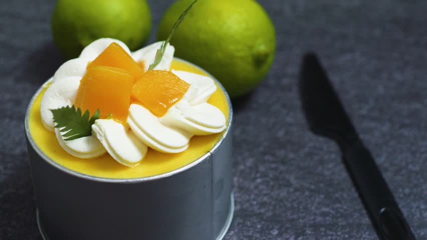Small mango mousse cake adorned with whipped cream rosettes, peach slices, and mint, resting on a dark textured surface beside limes and a knife, creating a vibrant scene