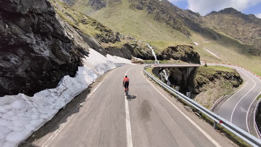 Epic shot of a cyclist conquering a challenging mountain pass. The road winds through stunning scenery, with snow patches and waterfalls adding to the dramatic landscape. Perfect for adventure, travel