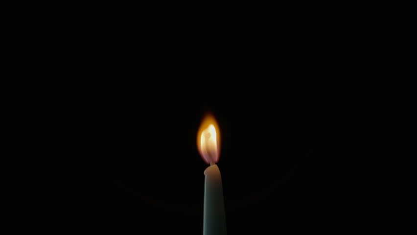 Tiny candle staying on black background in total stillness. Flickers move slightly across quiet walls of night. One steady flame giving feeling of warmth or devotion. Wax taper burning on dark surface