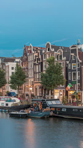 Panoramic timelapse hyperlapse of the illuminated famous Dancing Houses along the Amstel River day to night transition. Water channels with houseboats. Iconic landmark in Amsterdam, Netherlands