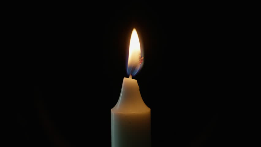 Glowing wax taper standing alone against shadowed backdrop closeup. Darkness fades under trembling flame inside quiet room. Burning white candle on black isolated background. Memory and church concept