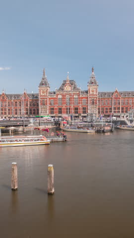 Timelapse of Amsterdam Central Railway Station front view with tour boats moored at the jetty, trams passing, and a canal in the foreground. Amsterdam, The Netherlands