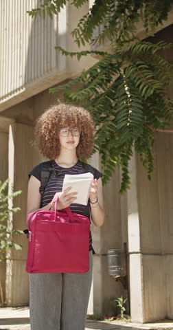 A thoughtful young female student with curly hair and glasses stands outdoors, holding notebooks and a red laptop bag. She appears to be contemplating her studies in an educational environment.
