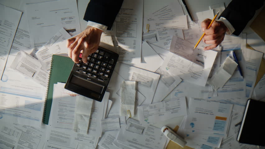 Accountant calculating corporate budget planning money profit at table top view. Business man hands holding documents analyzing expenses doing work using calculator over paper bills invoices indoors