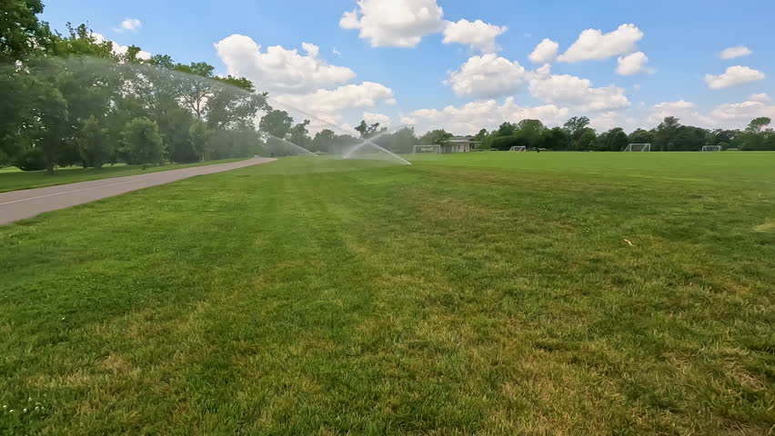 Sprinklers water grassy sports field at Forest Park in St. Louis, Missouri, USA. City park located in the heart of the city. Slow motion views, moving closer to the spraying water.