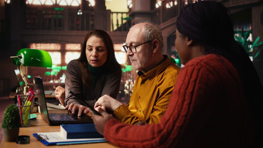Mature academic colleagues prepare documents for university coursework or exams, transcribing essential ideas from textbooks. Research and education in a quiet public library. Camera A.