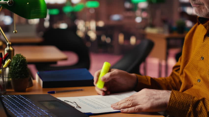 Elderly student using highlighter pen to memorize lesson papers, applying a study strategy with essential information from courses. Old man works on memorization for exams. Camera B.
