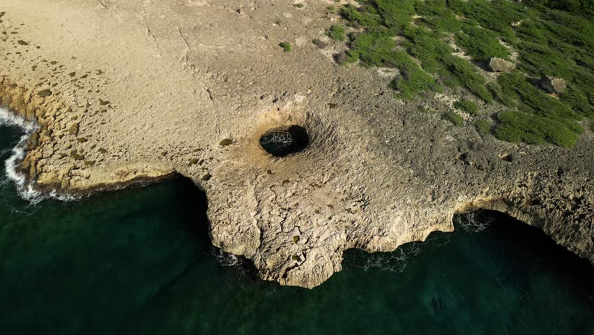Bright aerial over Watamula Hole, Curaçao: rugged coral‑rock coast, turquoise sea, lush greens and dramatic blowholes in vivid daylight—perfect for travel promos