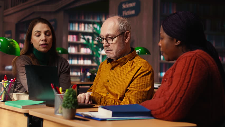 Mature diverse colleagues writing notes and ideas in campus study room, focusing on academic graduation preparation at the college library. Learning for personal development. Camera B.