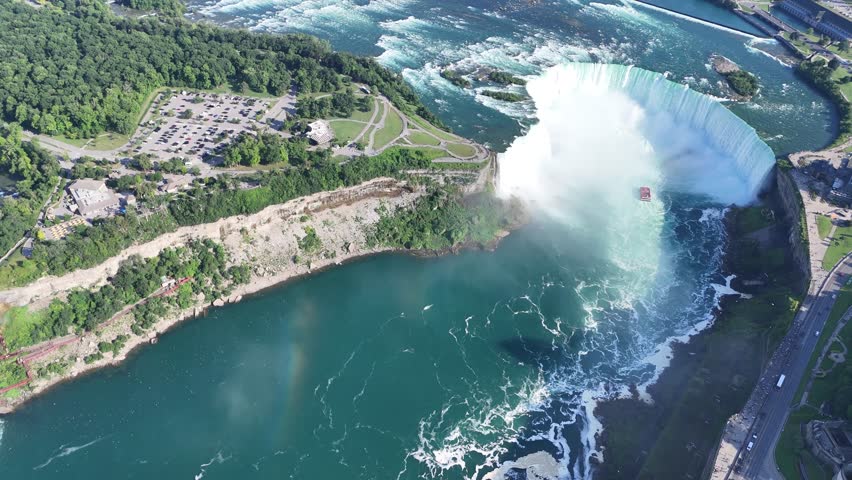 Drone flies over Horseshoe Falls capturing cruise ships below and a vivid rainbow in the mist, with a clear view of Niagara Falls State Park on the American side in the background