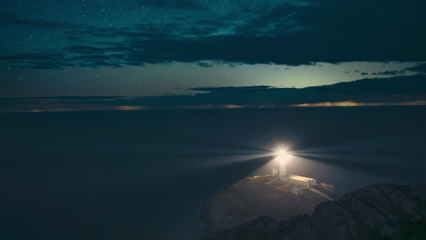 Timelapse of South Stack Lighthouse, Anglesey. A rotating beam cuts through cloudy night sky, stars, and sea. A stunning and atmospheric coastal scene of a Welsh maritime landmark.