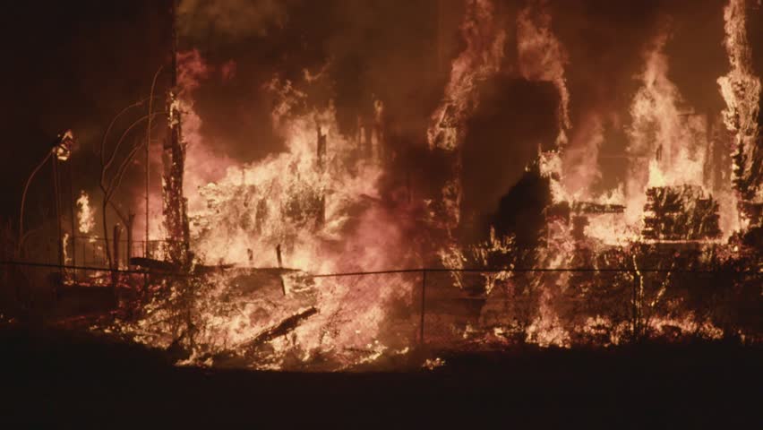 A wooden building collapses as towering flames and thick smoke rise into the night sky, captured from a safe distance behind a chain-link fence.